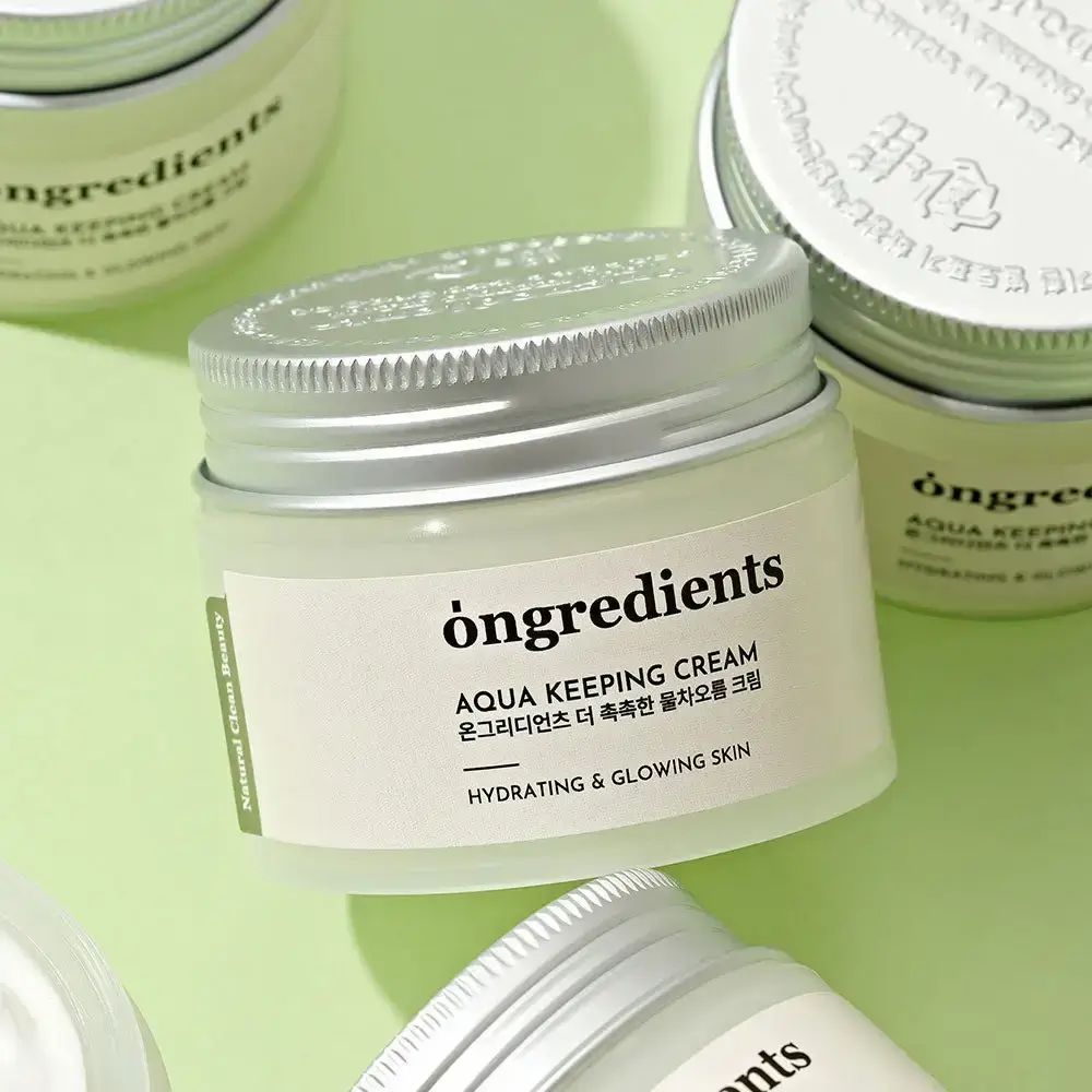 Creme-Glas mit silbernem Deckel. Aufschrift: òngredients, AQUA KEEPING CREAM, Hydrating & Glowing Skin. Mehrere Gläser auf grünem Grund.