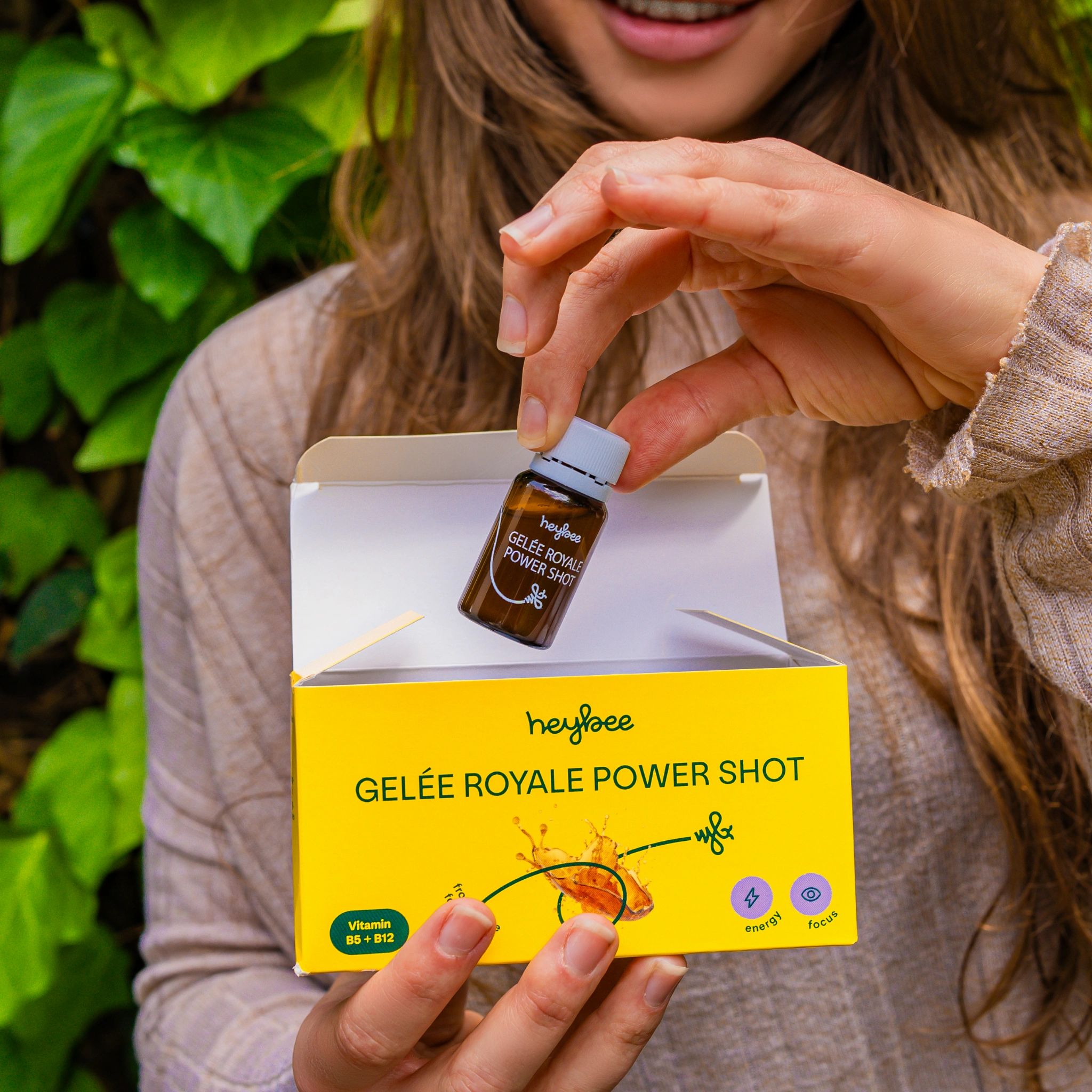 Frau hält Heybee Gelée Royale Power Shot. Gelbe Verpackung mit Produktinformationen. Enthält B5, B12 und C. Energie, Fokus.