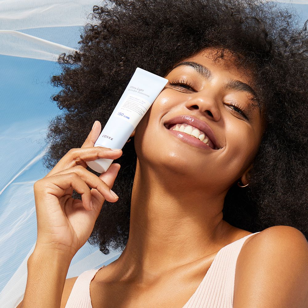 Frau hält COSRX Ultra Light Invisible Sunscreen Tube an ihre Wange.