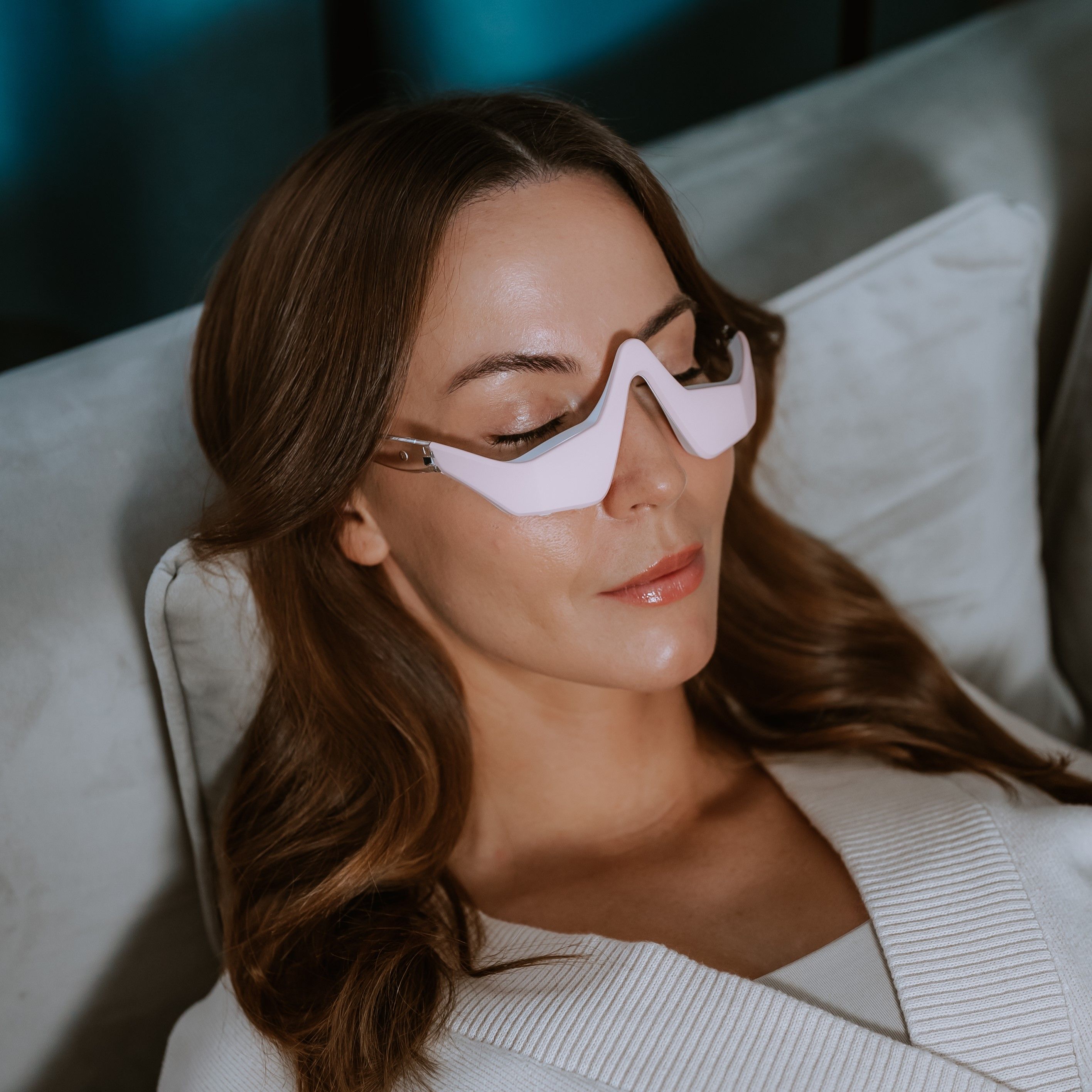 Frau liegt auf Sofa mit geschlossenen Augen, trägt rosa Augenmassagegerät in Brillenform.