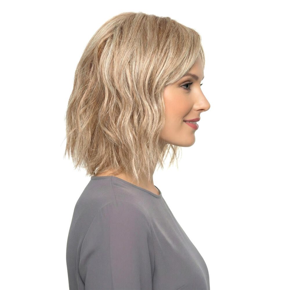 Blond gewellte Perücke mit Pony. Seitenansicht einer Frau mit grauer Bluse. Der Hintergrund ist weiß.