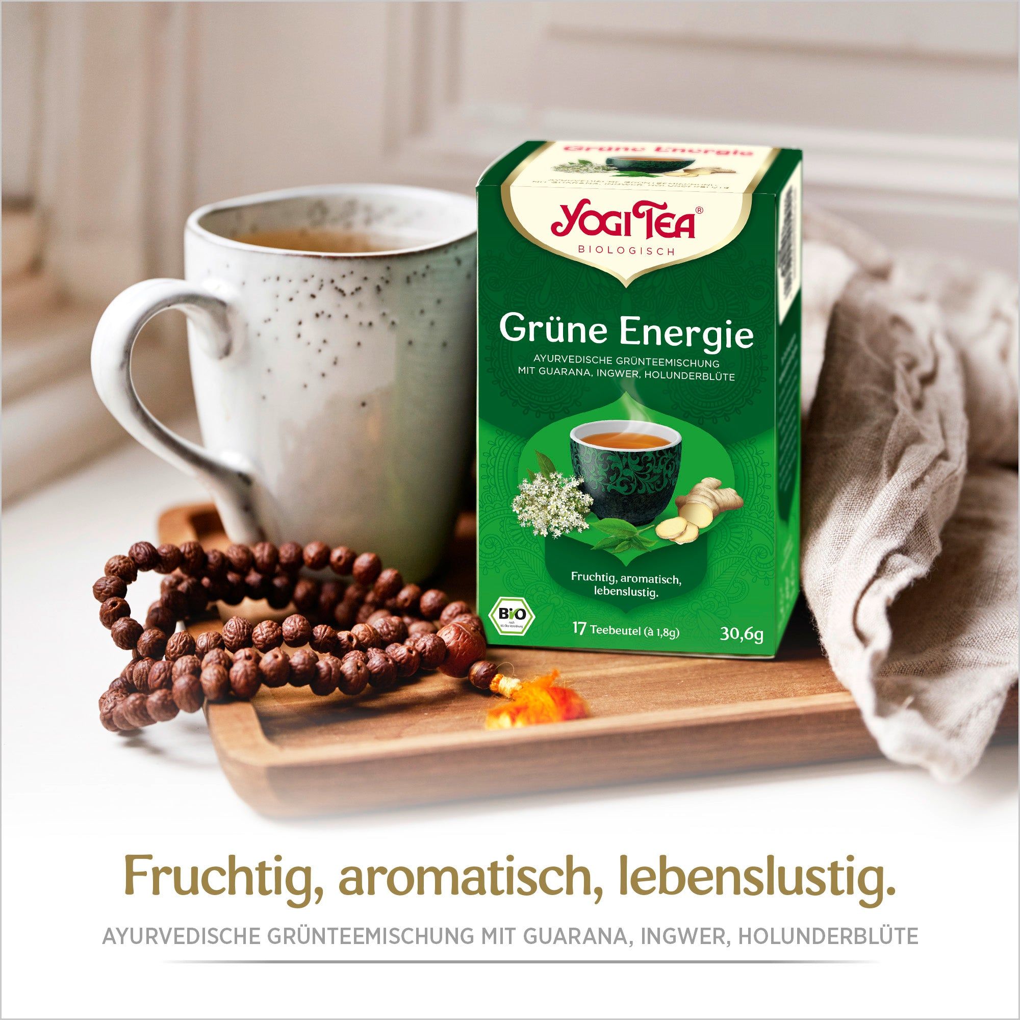 Eine grüne Tee-Schachtel, Yogi Tea, Grüne Energie, neben einer Tasse Tee und einer Gebetskette. Text und Illustrationen von Kräutern.