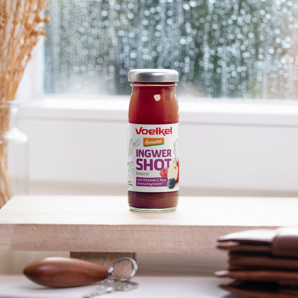 Glasflasche Voelkel Ingwer Shot Beere. Aufschrift: Ingwer Shot Beere. Auf Holzoberfläche. Hintergrund: Fenster mit Regentropfen.