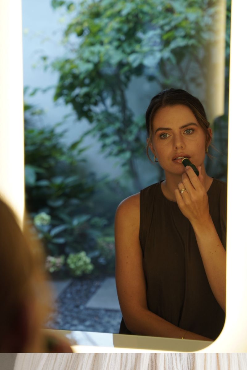 Frau im Spiegel, trägt Lippenbalsam auf. Spiegel mit hellem Rahmen, Garten im Hintergrund.