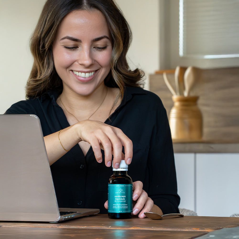 Frau mit Laptop und Flasche. Sie hält eine Flasche Pure Focus Flüssigextrakt. Löffel auf dem Tisch.