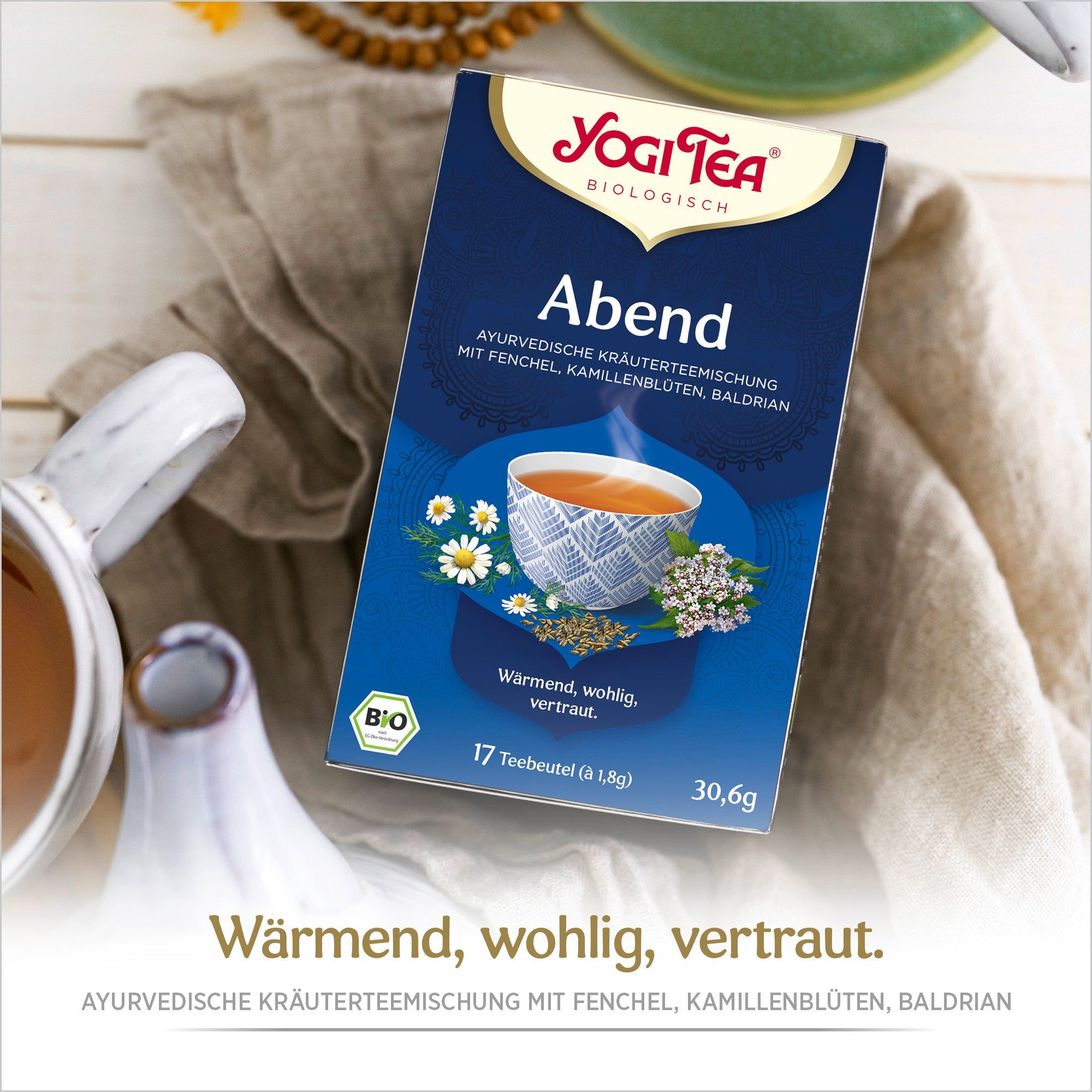 Blaue Teepackung mit Logo und Produktbezeichnung. Bio-Siegel. Tasse Tee auf Tischdecke. Text: Warm, wohlig, vertraut.