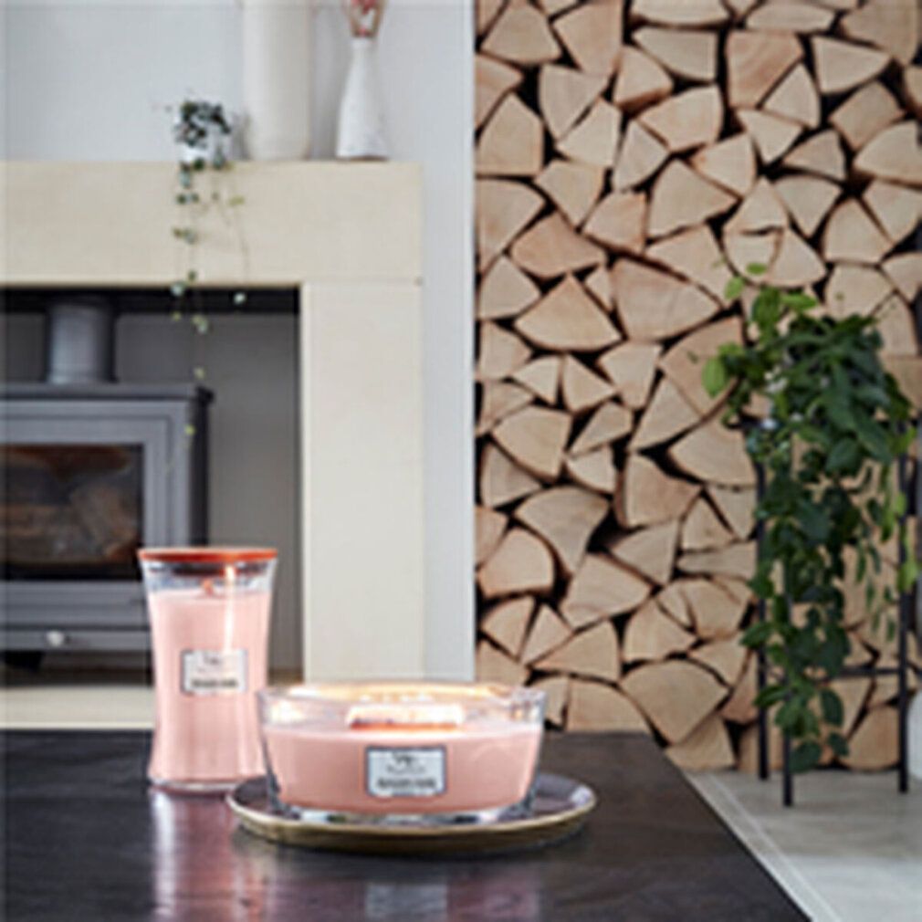Zwei Kerzen, eine hoch, eine flach, auf einem Tisch. Holzstapel im Hintergrund. Kerzen mit Logo und Produktnamen.