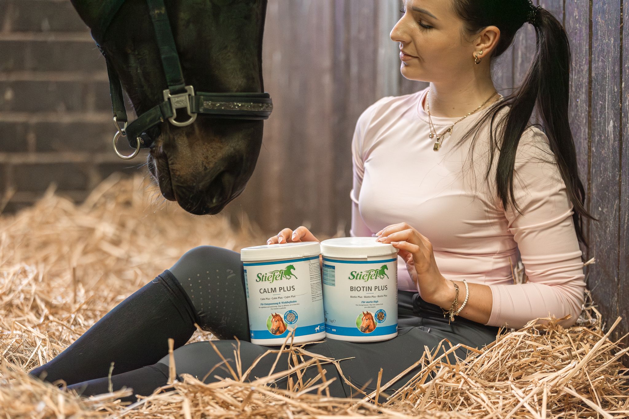 Frau sitzt im Heu und hält zwei weiße Behälter. Aufschrift: Stiefel Calm Plus und Stiefel Biotin Plus. Pferd im Hintergrund.