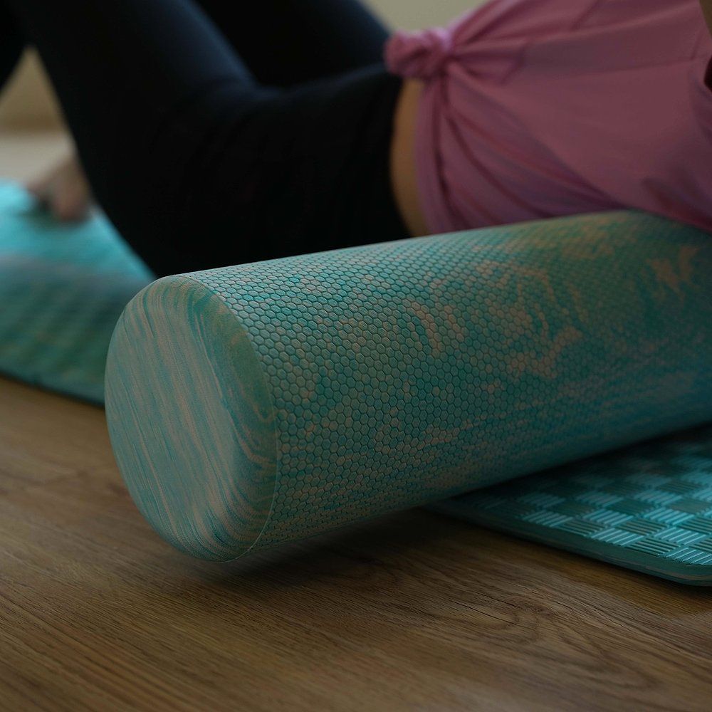 Zylindrischer Pilates-Roller, blau-weiß marmoriert, auf grüner Matte. Person liegt mit Bein auf dem Roller.