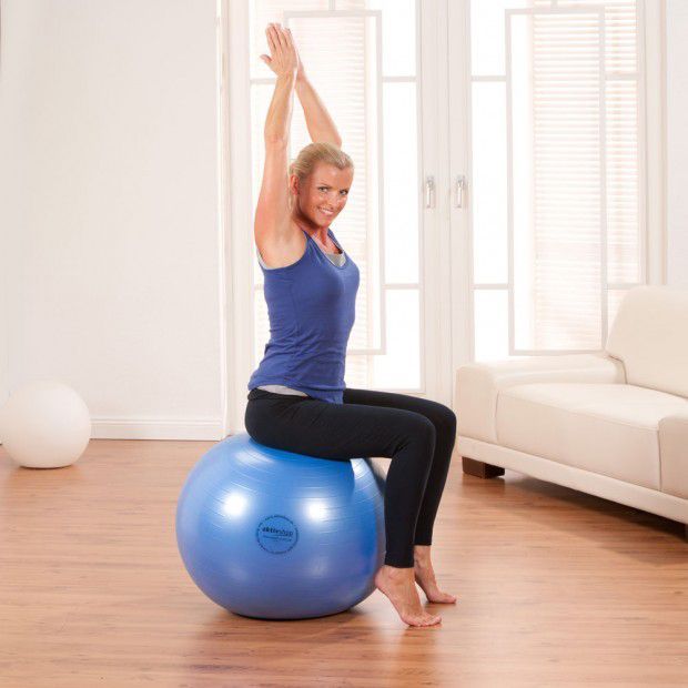 Frau sitzt auf blauem Fitnessball, Arme nach oben gestreckt. Helle Umgebung.