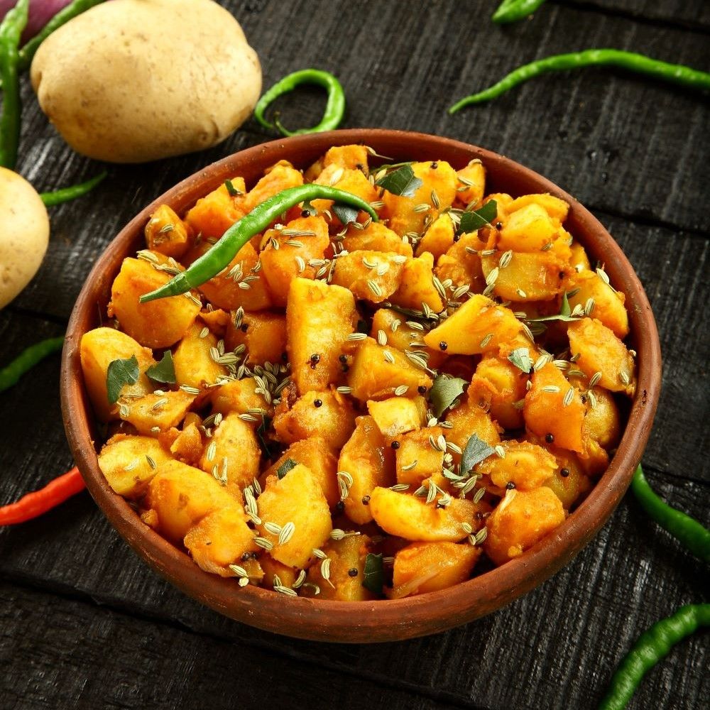 Schüssel mit gebratenen Kartoffeln, Gewürzen und grünen Chilis. Auf dunklem Holz. Kartoffeln und Chilis im Hintergrund.