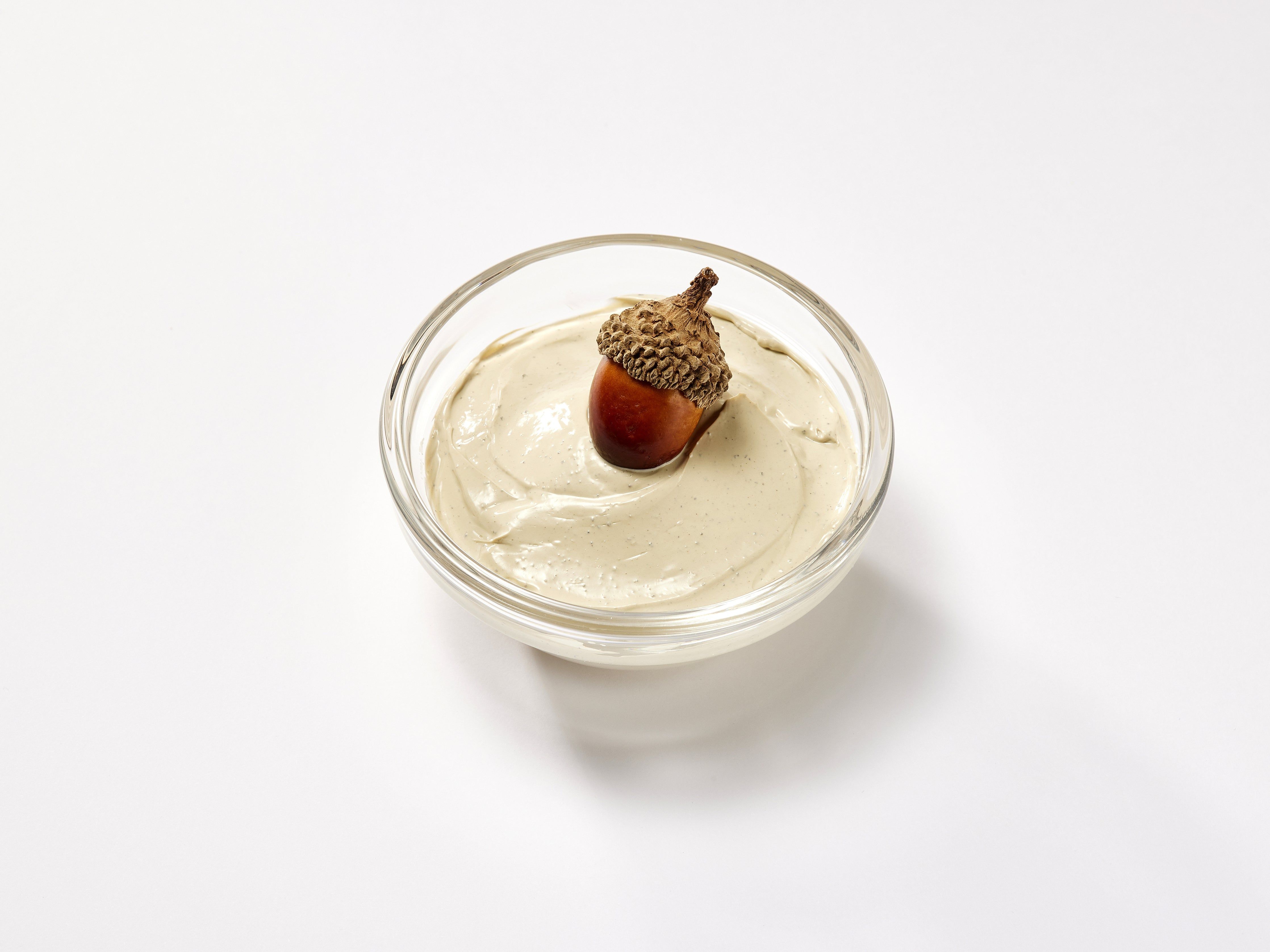 Klare Glasschale mit cremefarbener Substanz und Eichel. Auf weißem Hintergrund.