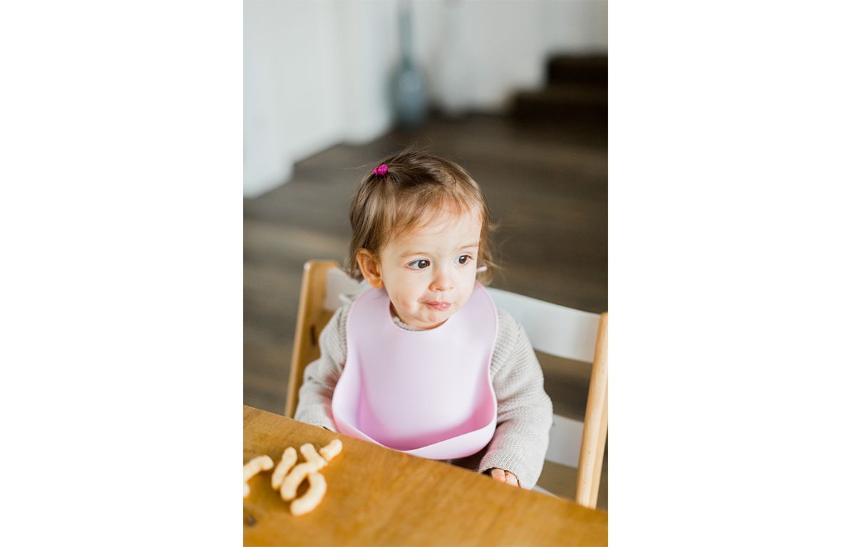 Baby mit zartrosa Silikonlätzchen am Tisch. Kind sitzt in einem Hochstuhl.
