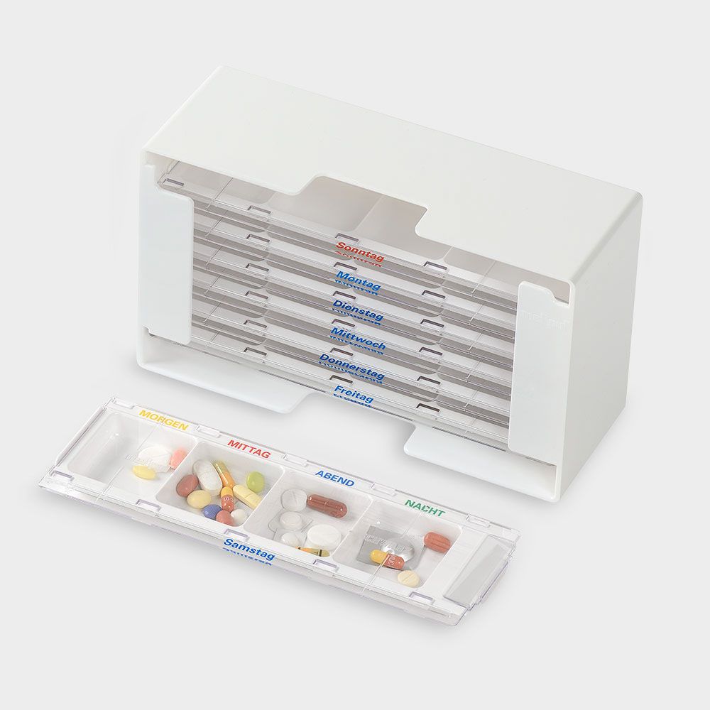 Weißer Medikamenten-Dosierer mit 7 Tages- und 4 Fächern. Ein Fach ist herausgenommen und mit Tabletten gefüllt. Beschriftung in Deutsch.