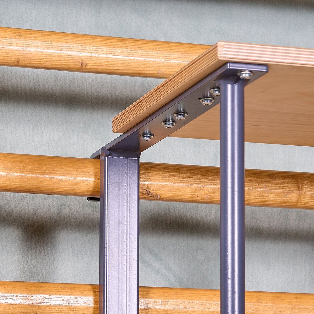 Nahaufnahme des Sprossenwandsitzes. Holzplatte mit Metallrahmen, verschraubt. Rahmen an Sprossenwand befestigt. Grauer Metallrahmen.