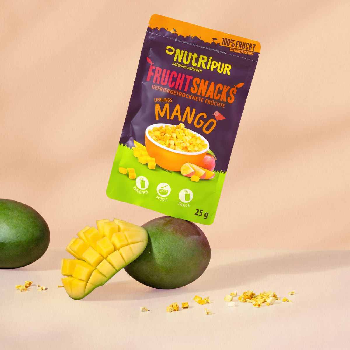 Tüte gefriergetrockneter Mango-Fruchtsnacks, daneben Mango und Mangostückchen. 25g.