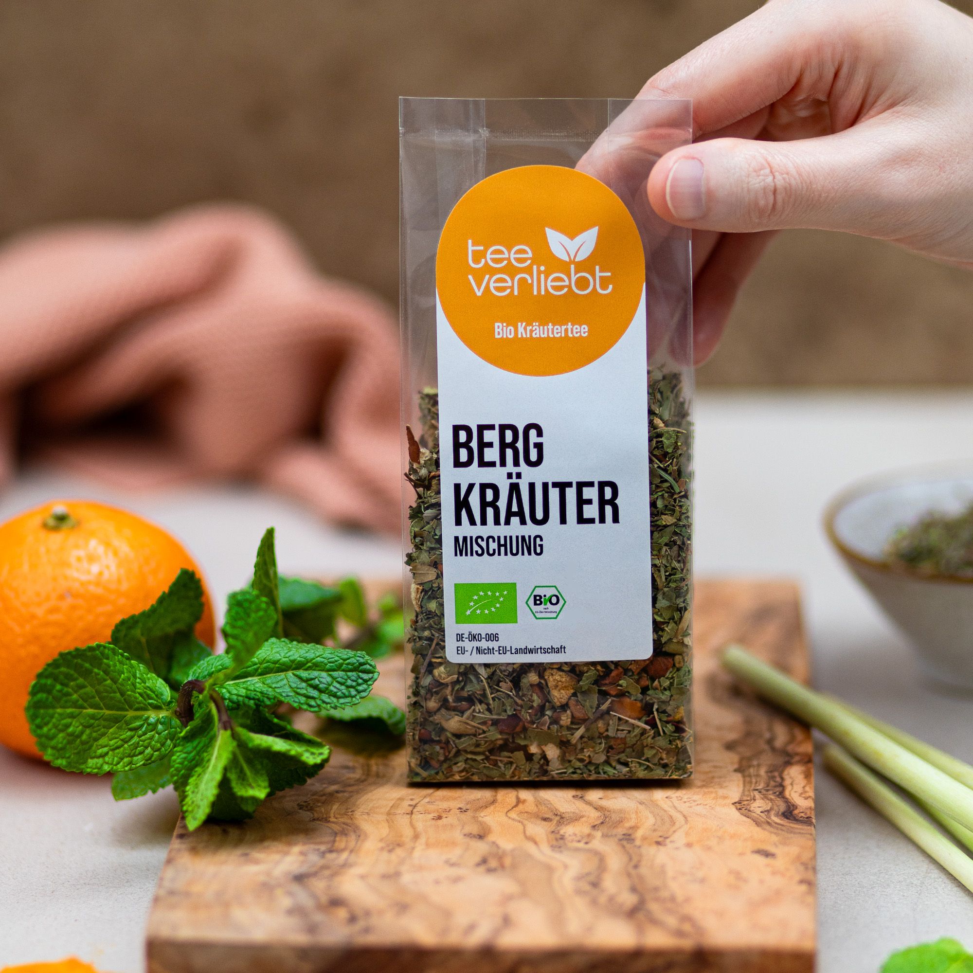 Hand hält transparente Verpackung mit Kräutermischung. Aufkleber "tee verliebt" und "Bio Kräutertee". Text: "Berg Kräuter Mischung". Bio-Siegel. Orange, Minze.