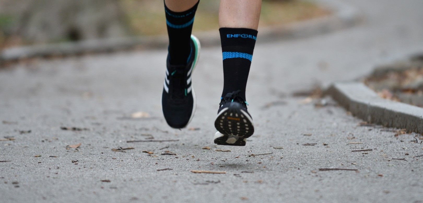 Una persona corre su una strada, indossando calzini neri con strisce blu e scarpe sportive. I calzini sono Enforma Achilles Support. L'attenzione è sui piedi.
