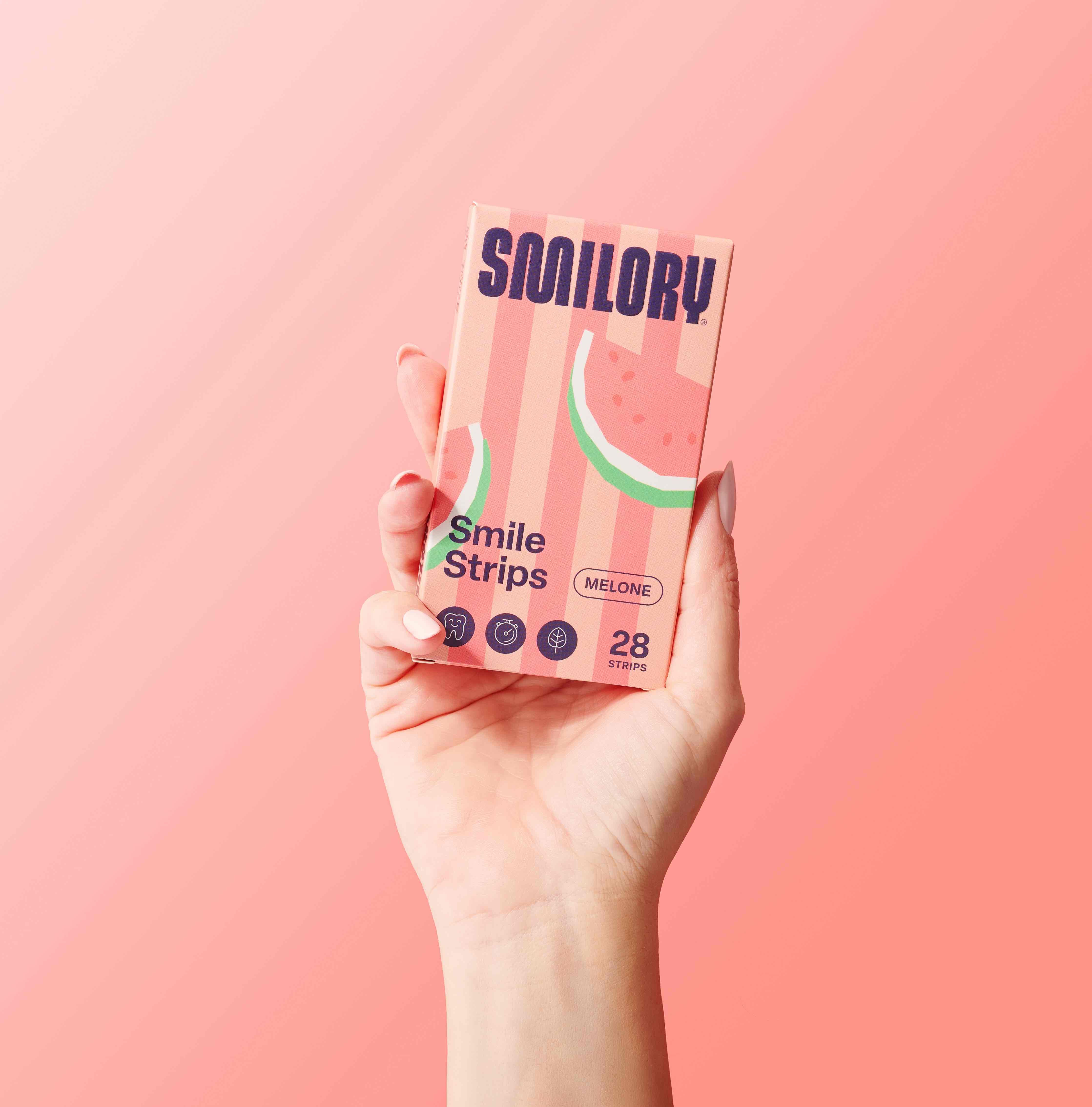 Hand hält eine Packung Smilory® Smile Strips. Rosa gestreifte Verpackung mit Wassermelonen-Illustration. Text: Smile Strips, Melone, 28 Strips.