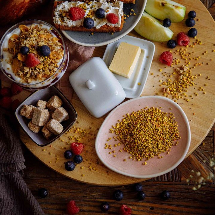 Draufsicht auf Frühstück mit Blütenpollen. Enthält Müsli, Brot mit Beeren, Butter, Zuckerwürfel, Obst und Blütenpollen.