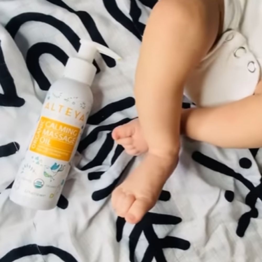 Weiße Flasche mit Pumpe neben Babyfüßen. Aufschrift: Alteya Organics, Calming Massage Oil. Auf Decke.