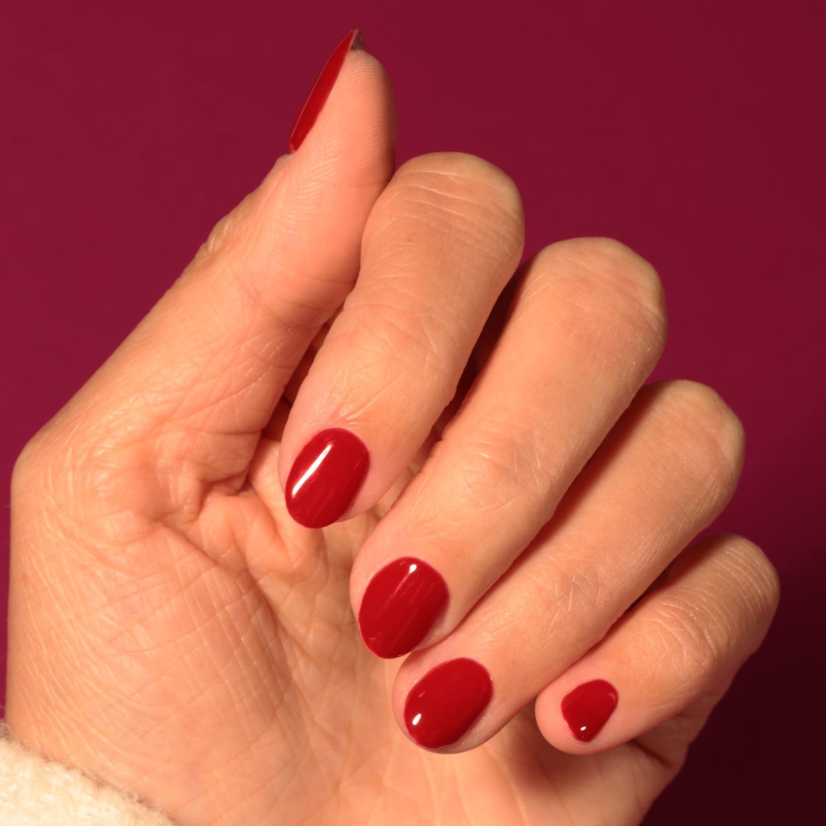 Hand mit lackierten, roten Nägeln. Die Nägel sind kurz und glänzend. Der Hintergrund ist rosa.