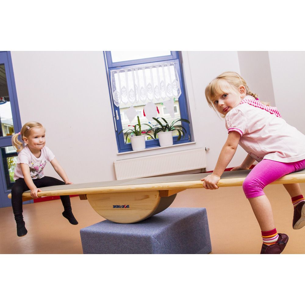 Kinder balancieren auf einem Holz-Element. Darunter ein graues Polster. Im Hintergrund Fenster und weitere Kinder.