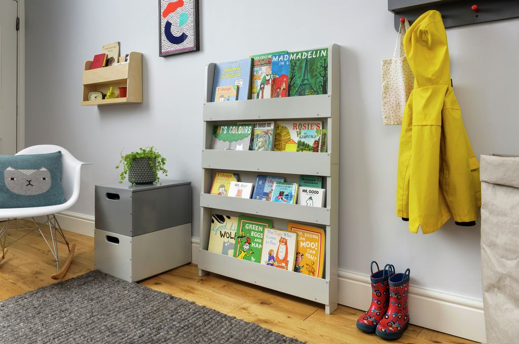 Hellgraues Bücherregal mit Büchern in einem Raum. Bücher mit farbigen Einbänden. Der Raum hat eine graue Wand, einen Teppich und einen Stuhl.