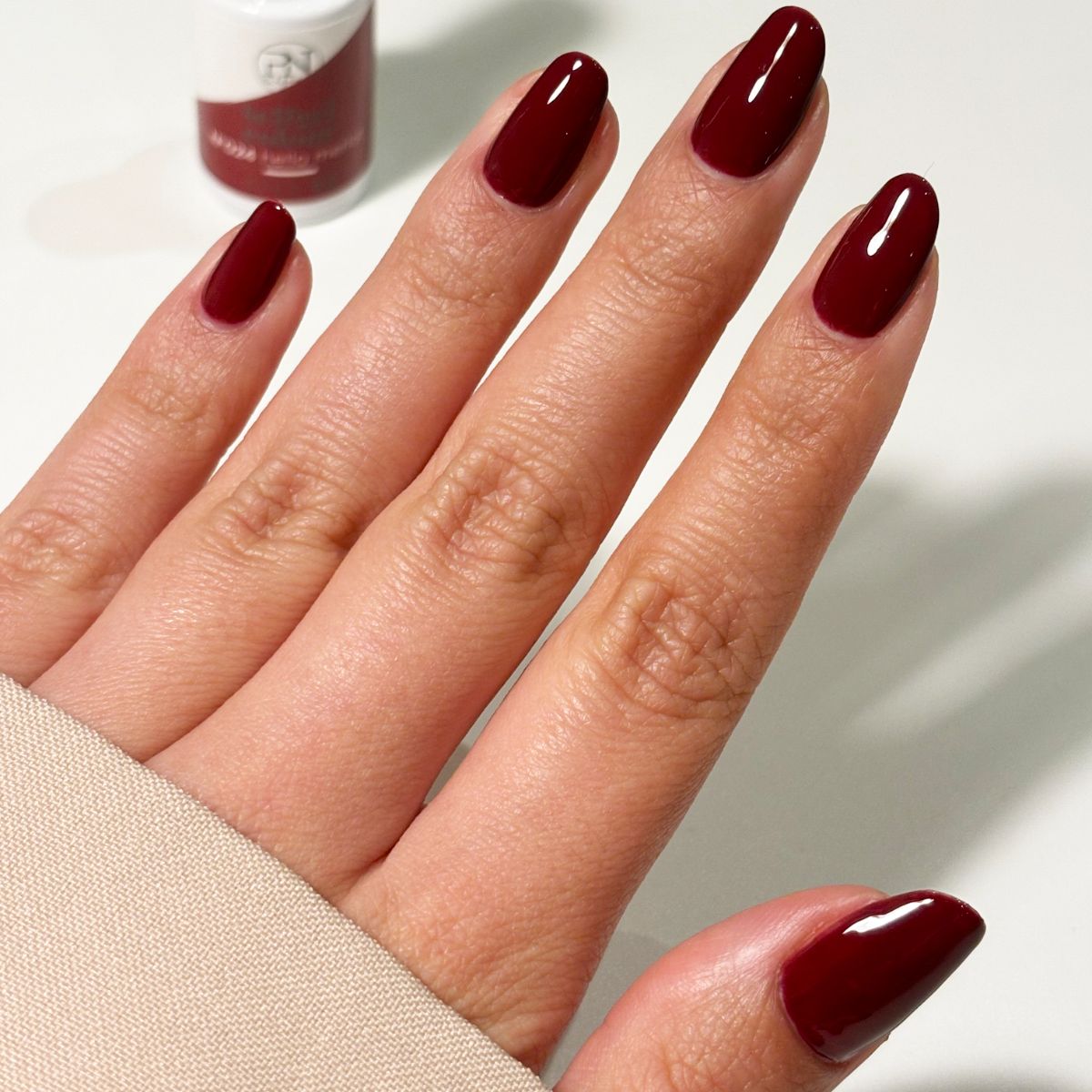 Hand mit lackierten Nägeln in Burgunderrot. Ein Nagellackfläschchen im Hintergrund. Nägel sind oval geformt.