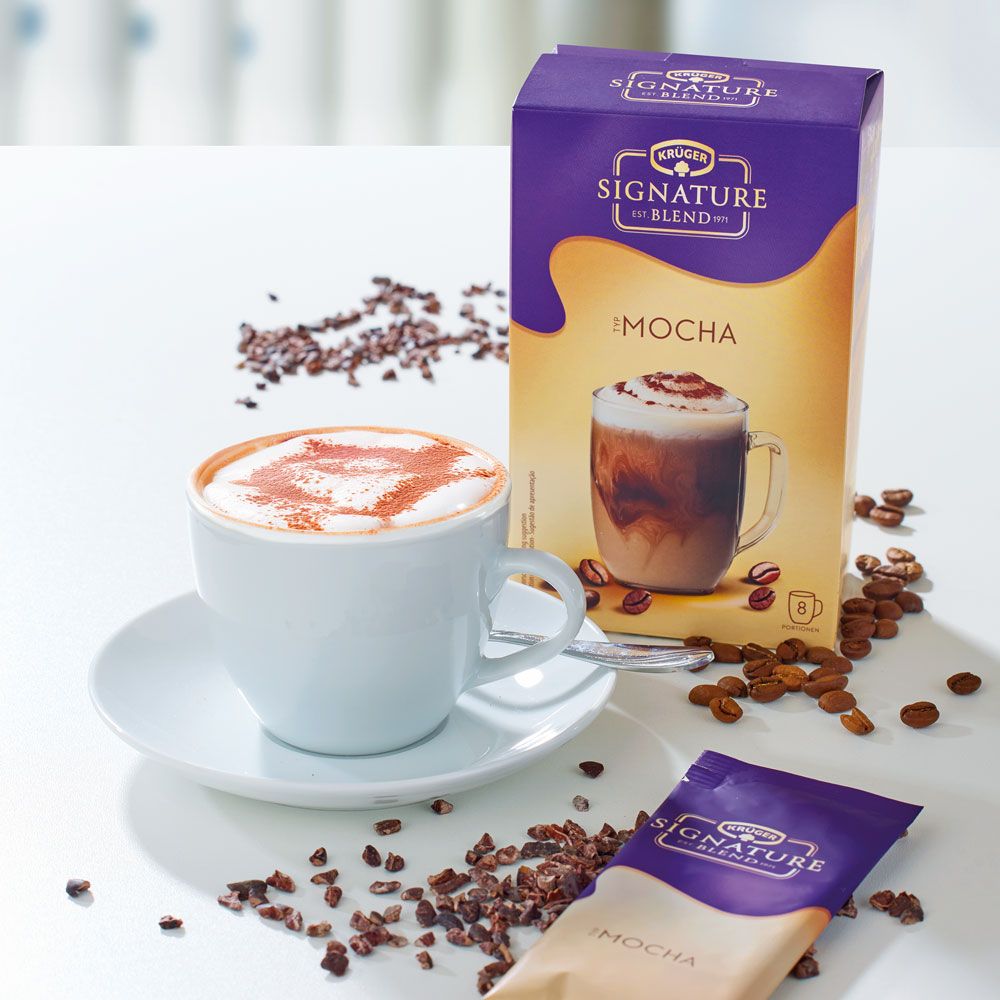 Produktverpackung und Getränk in Tasse. Aufschrift: Krüger Signature Blend Mocha. Dazu Kakaobohnen und ein Beutel.