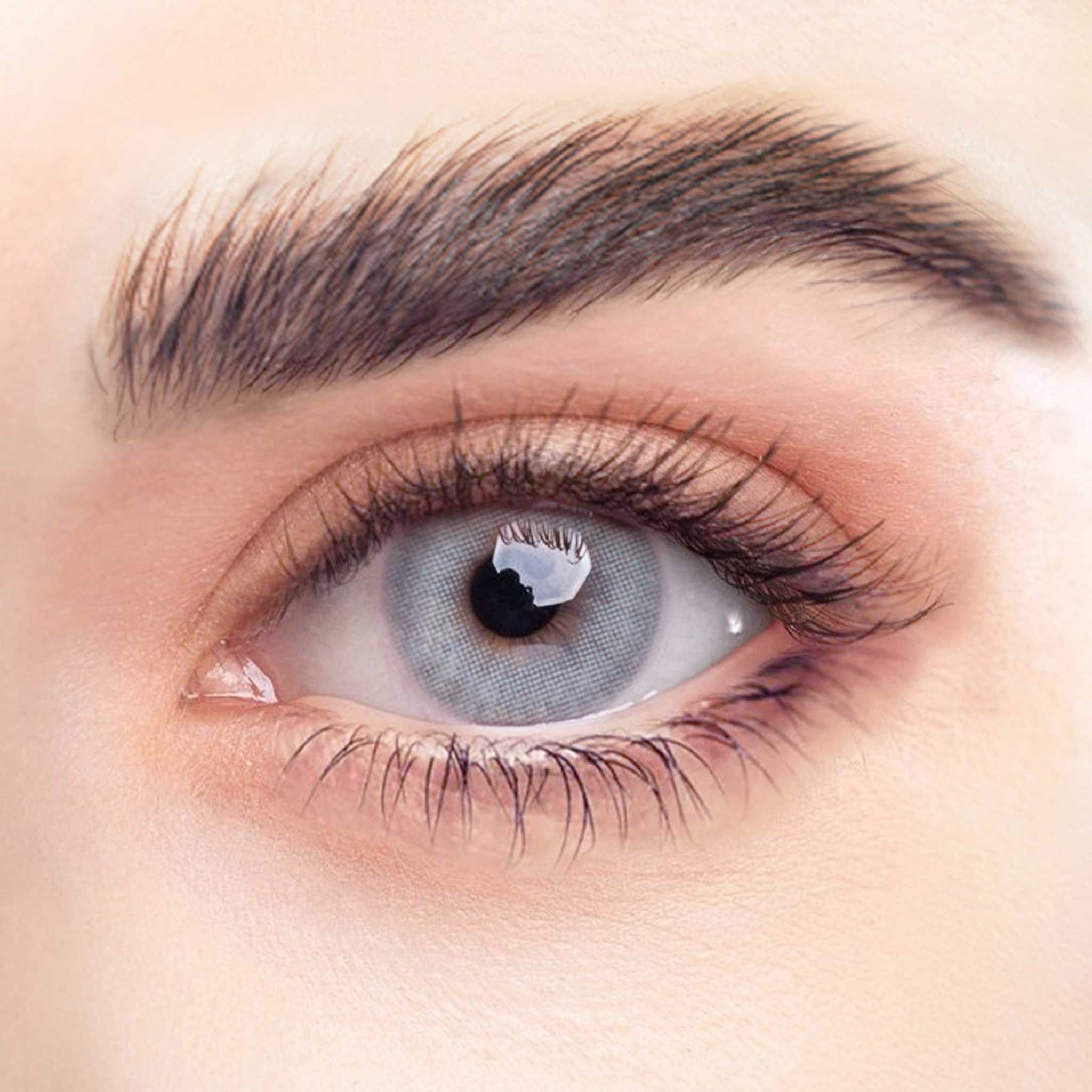 Nahaufnahme eines Auges mit grauer Kontaktlinse. Sichtbar sind Wimpern, Augenbraue und Haut. Das Auge reflektiert Licht.