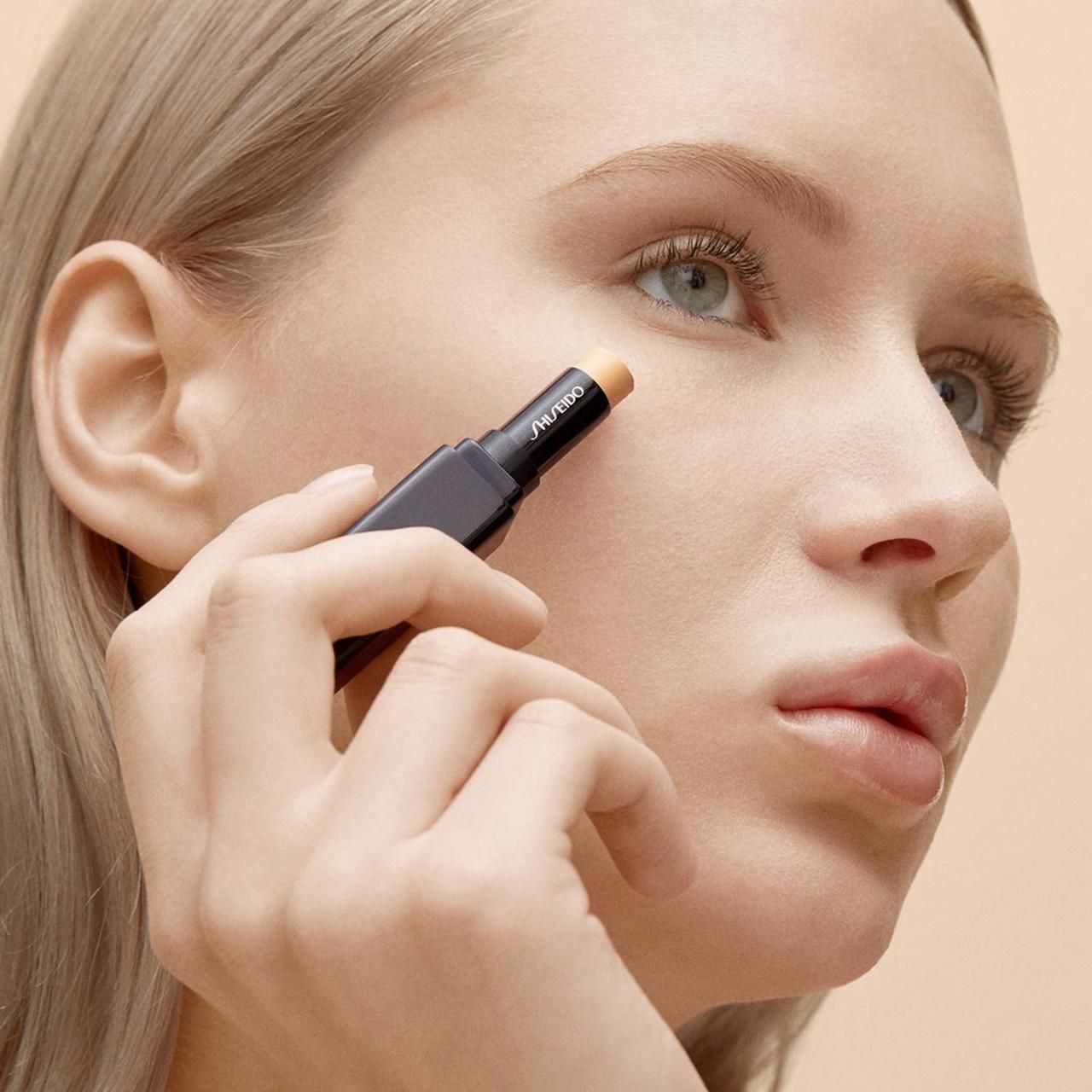 Frau trägt Concealer-Stick unter dem Auge auf. Schwarzer Stick, Marke Shiseido.