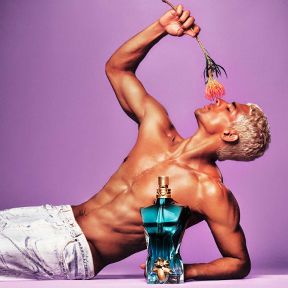 Mann mit blonden Haaren, hält Blume. Jean Paul Gaultier "Le Beau" Flakon in Torsoform. Auf lila Hintergrund.