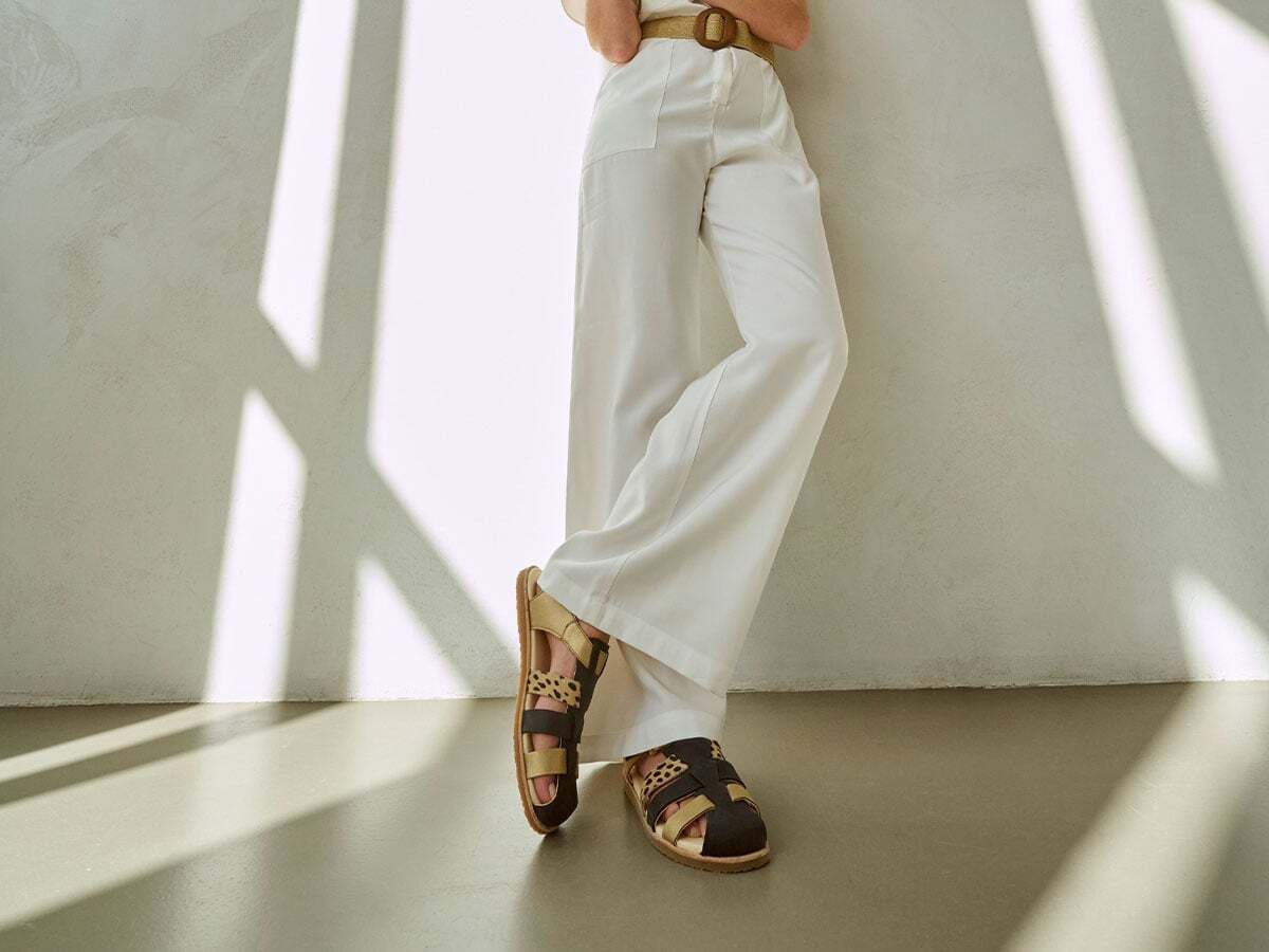 Person trägt Sandalen mit Riemen in Schwarz, Gold und Leopardenmuster. Offene Zehenpartie. Marke BÄR.