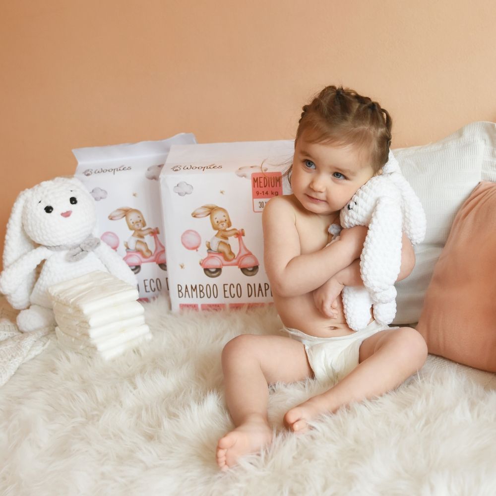 Baby sitzt auf Bett, hält Stofftier-Hase. Im Hintergrund Windelpackungen.