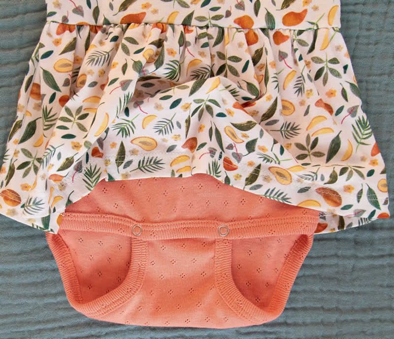 Nahaufnahme Bio-BW Mango Kleid. Weißes Oberteil mit Fruchtmuster, orangefarbener Unterteil. Kurze Ärmel, Rüschenrock. Lochmuster am Unterteil.