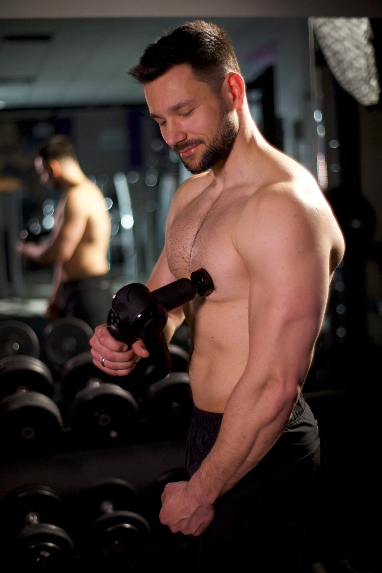 Mann in einem Fitnessstudio, der eine schwarze Massagepistole auf seiner Brust verwendet. Er steht vor Gewichten.