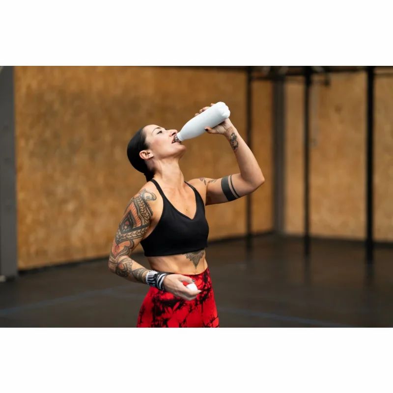 Frau trinkt aus einer weißen Trinkflasche. Sie trägt Sportkleidung und hat Tattoos.