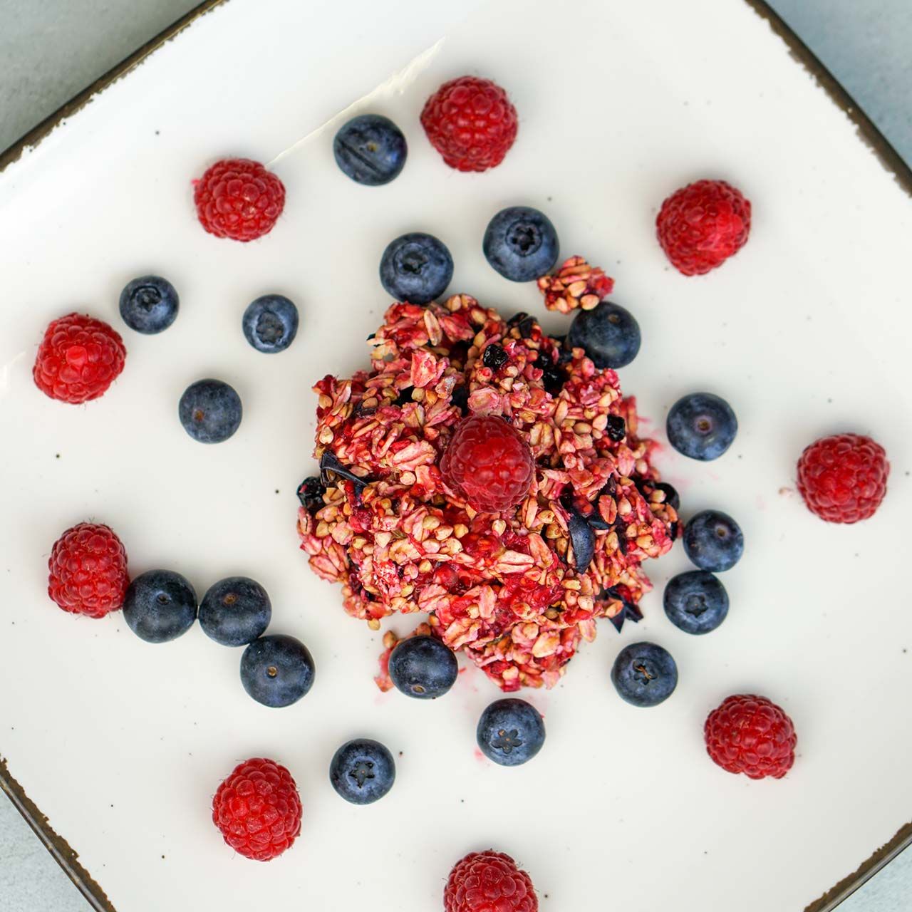 Müsli mit Himbeeren und Blaubeeren auf einem weißen Teller. Himbeeren und Blaubeeren umgeben das Müsli.