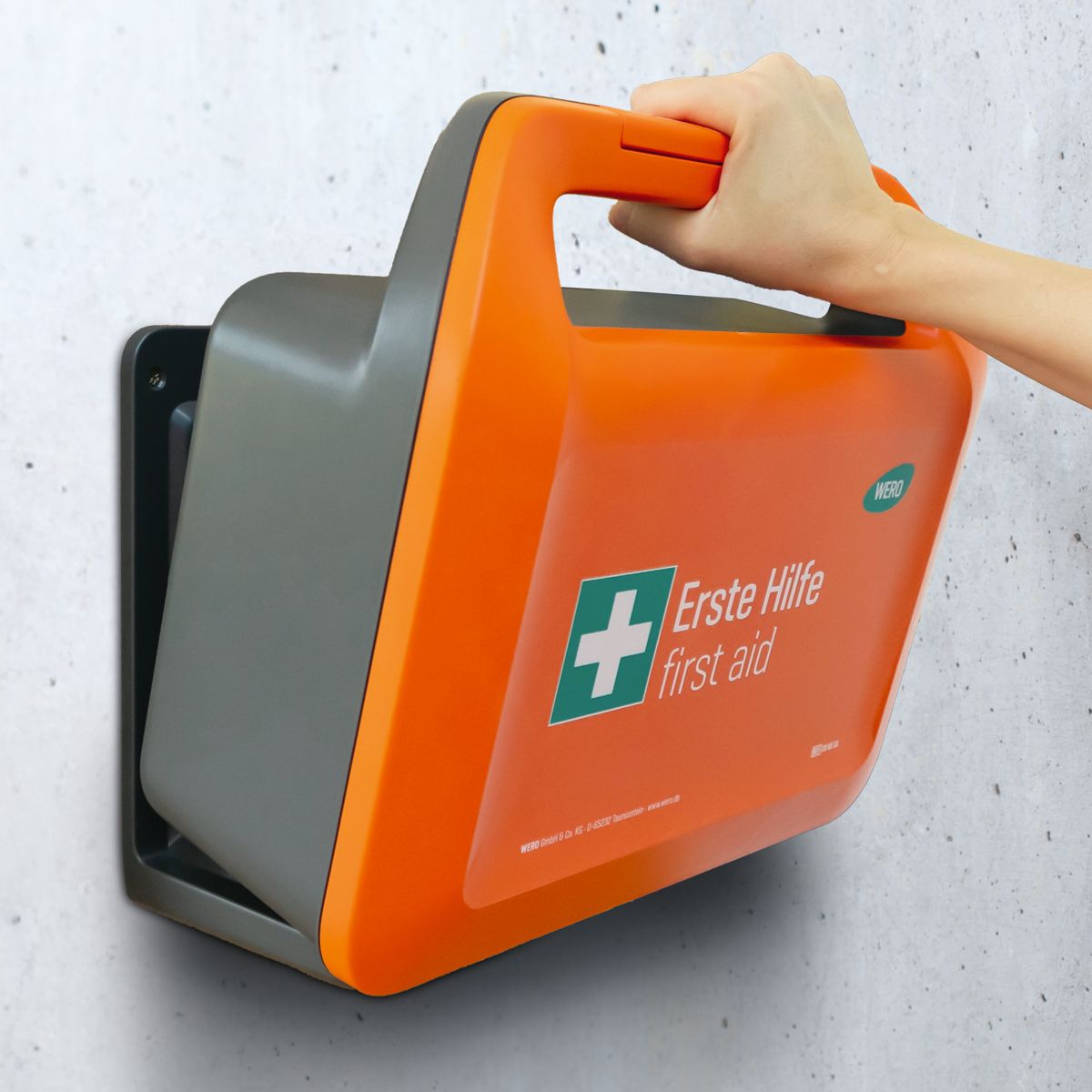 Orange Erste-Hilfe-Koffer an Wandhalterung. Hand greift nach Griff. Text: Erste Hilfe, Food processing. WERO Logo.