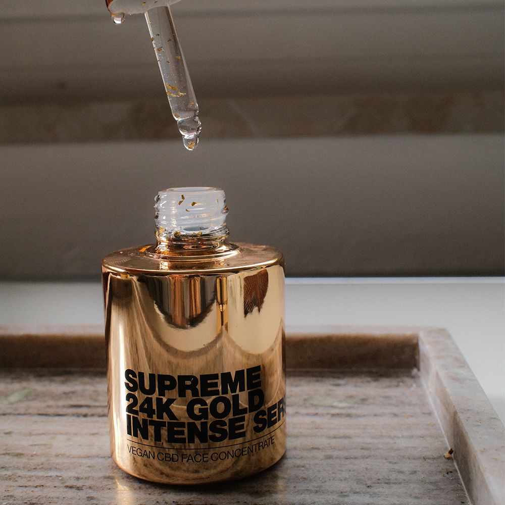 Goldene Serumflasche mit Pipette. Tropfen fallen in die Flasche. Aufschrift: Supreme 24K Gold Intense Serum.