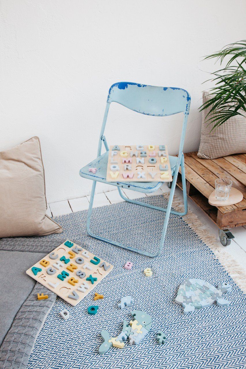 Zwei Holzpuzzles mit Buchstaben auf einem Teppich. Ein blaues Klappstuhl und Dekorationen im Hintergrund.