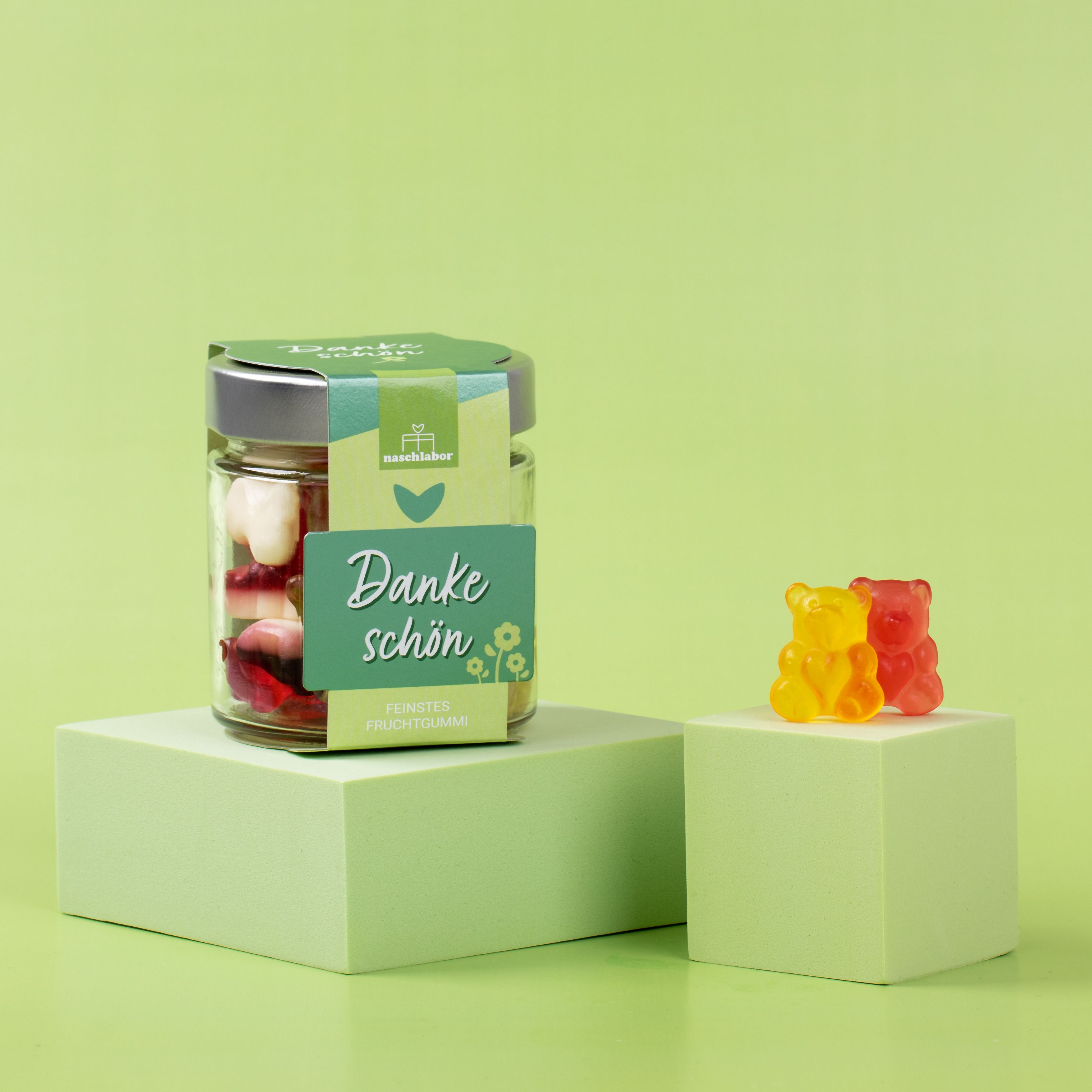 Glas mit Fruchtgummi auf Podest. Grün-weißes Etikett mit "Danke schön" und Logo. Zwei Fruchtgummi-Bären daneben auf Podest.