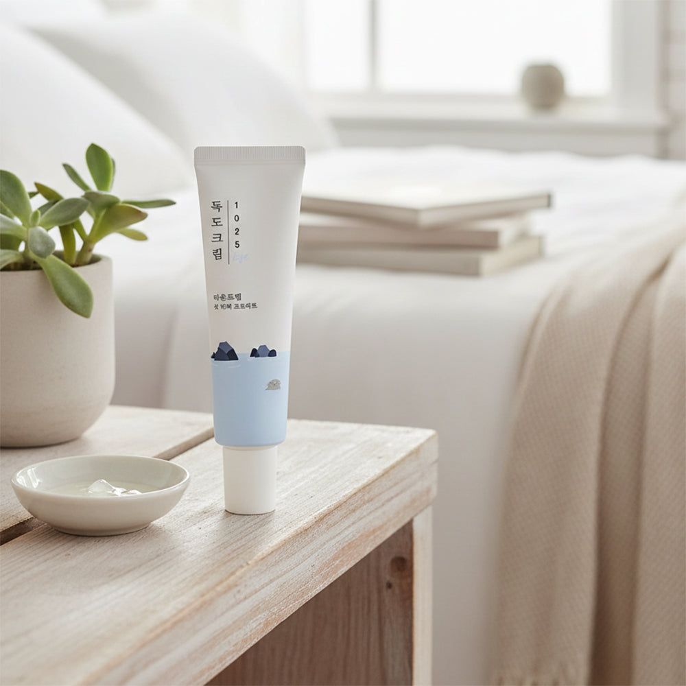 Weiße Tube mit blauer Aufschrift und Illustration. Steht auf einem Nachttisch neben einer Schale mit Creme und einer Pflanze.