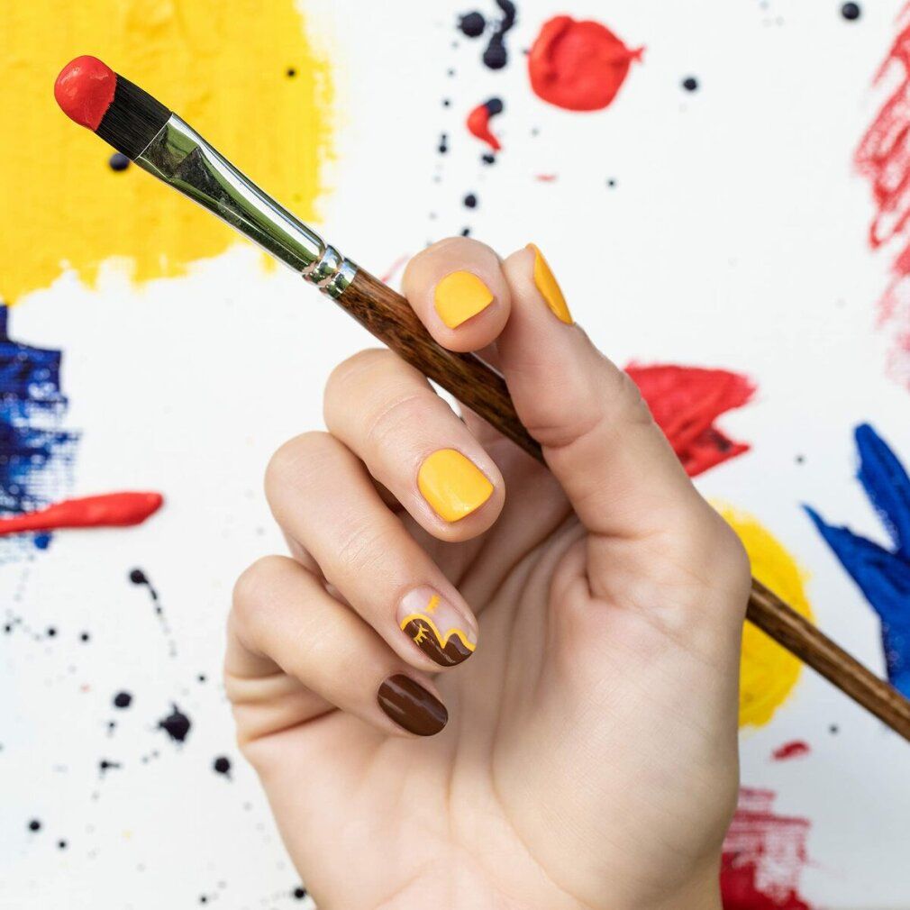 Hand mit gelb und braun lackierten Nägeln. Pinsel mit roter Farbe. Hintergrund mit bunten Farbspritzern.