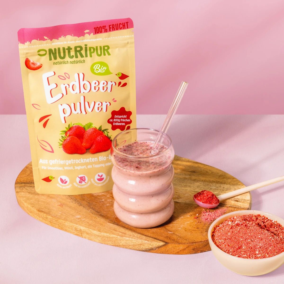 Produktpräsentation: Erdbeerpulver, Glas mit Erdbeershake, Löffel mit Pulver, Schale mit Pulver. Auf Holzbrett.