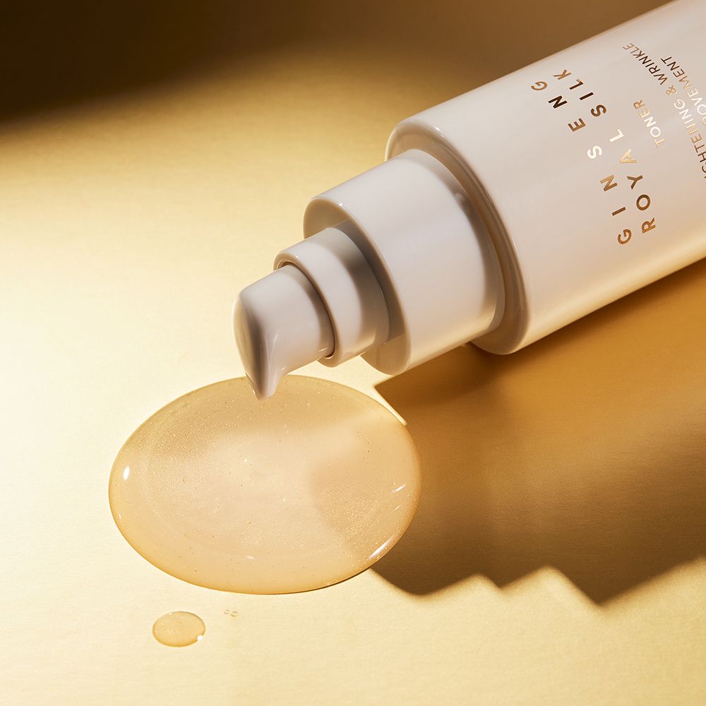 Beige Flasche, aus der Flüssigkeit tropft. Aufschrift: Ginseng Royal Silk. Flüssigkeit auf goldfarbenem Untergrund.