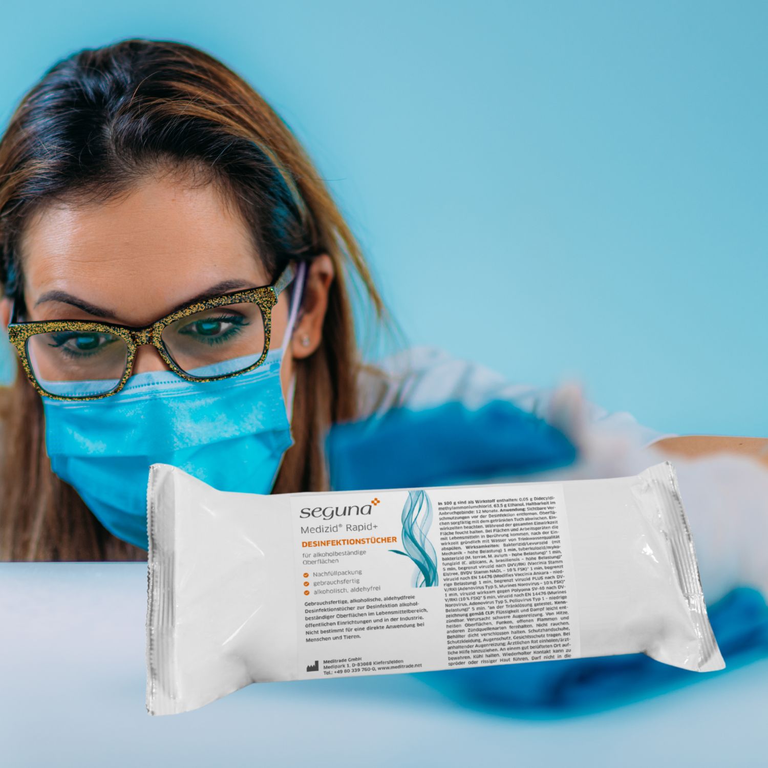 Person mit Brille und Maske hält eine Packung SEGUNA Medizid Rapid+ Desinfektionstücher. Produktinformationen und Logo sichtbar.