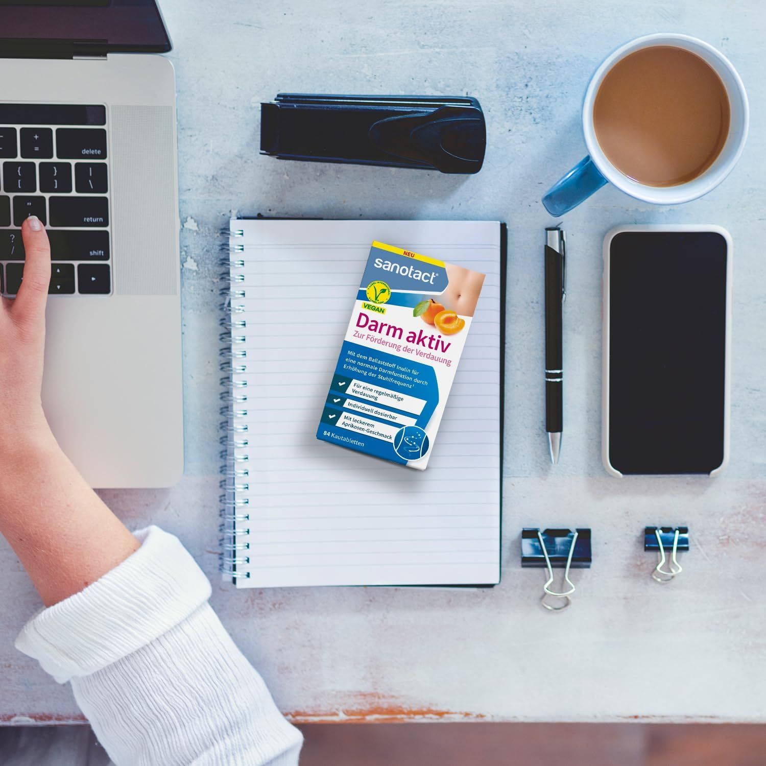Sanotact Darm aktiv Kautabletten. Verpackung auf Schreibtisch mit Laptop, Notizbuch und Smartphone.