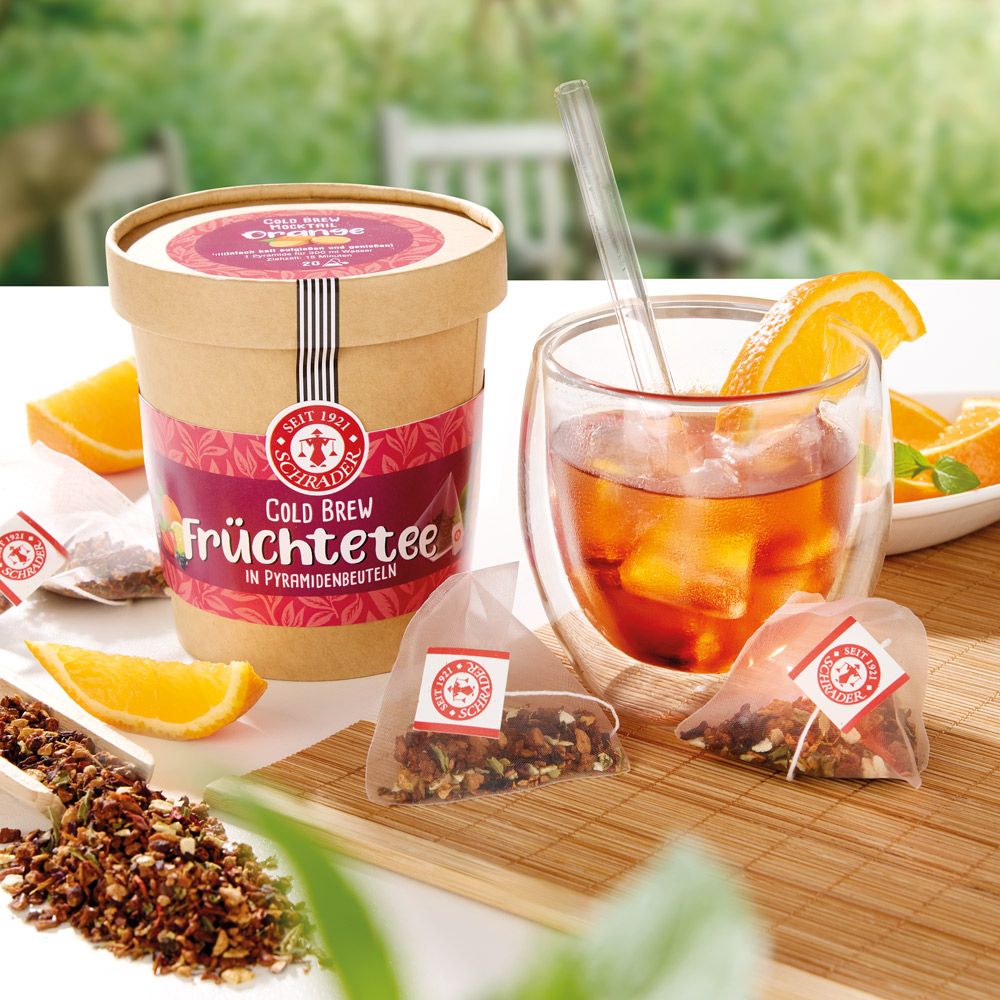 Pappdose mit Deckel, Glas mit Getränk und Orangenscheibe, Teebeutel, Orangenscheiben. Aufschrift: Schrader Cold Brew Orange.